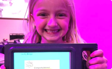 A smiling child holding an electronic bingo device that displays a 'Congratulations!' message, with a purple background.