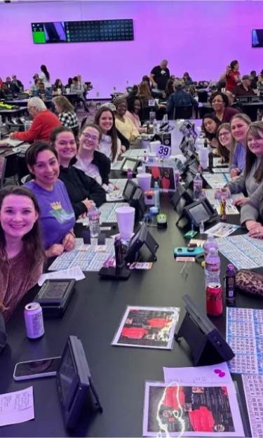 A group of people at a bingo hall, smiling with bingo cards and devices on the table.